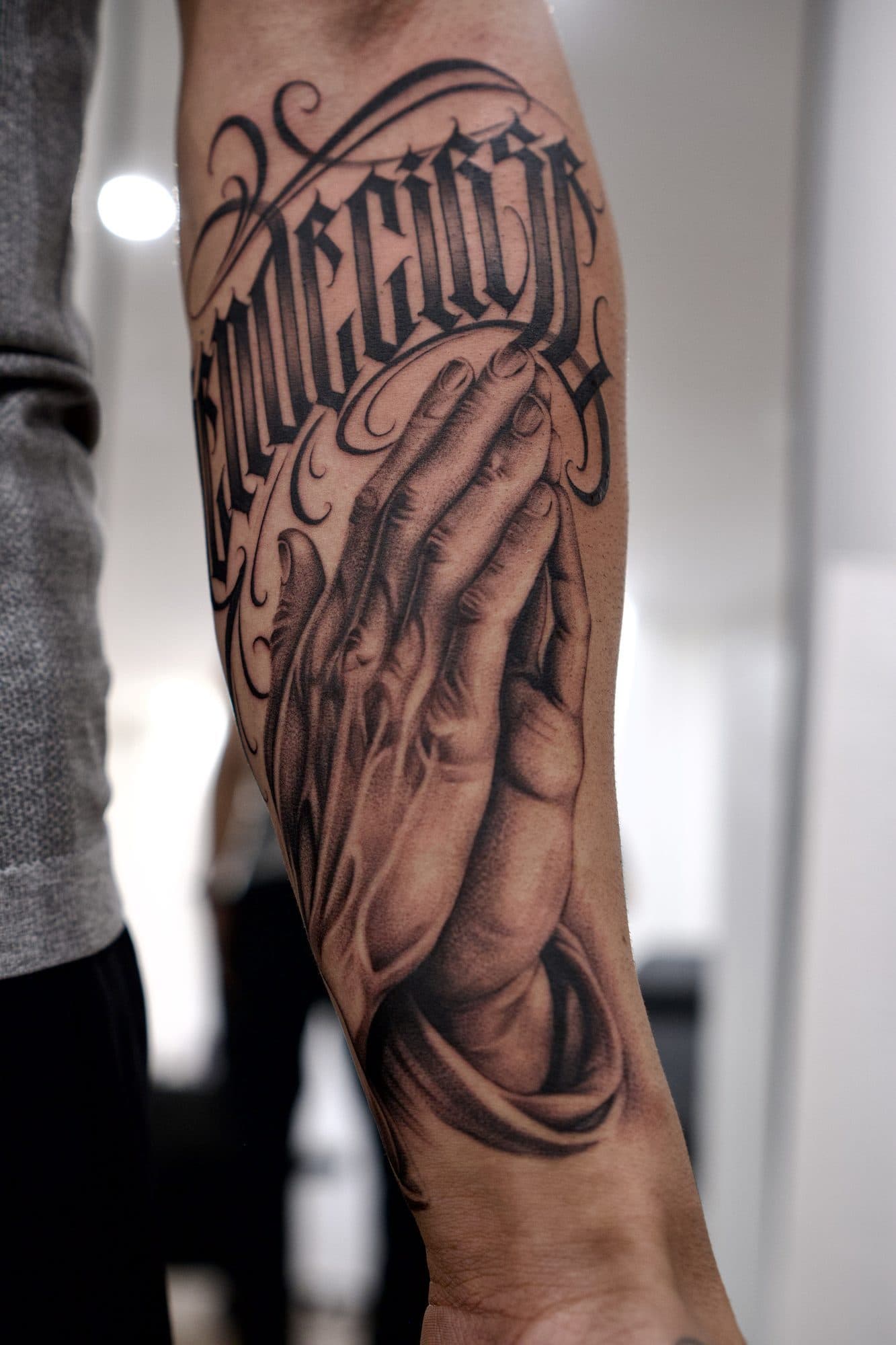 Antebraço em black and grey com mãos em oração sob lettering em blackletter