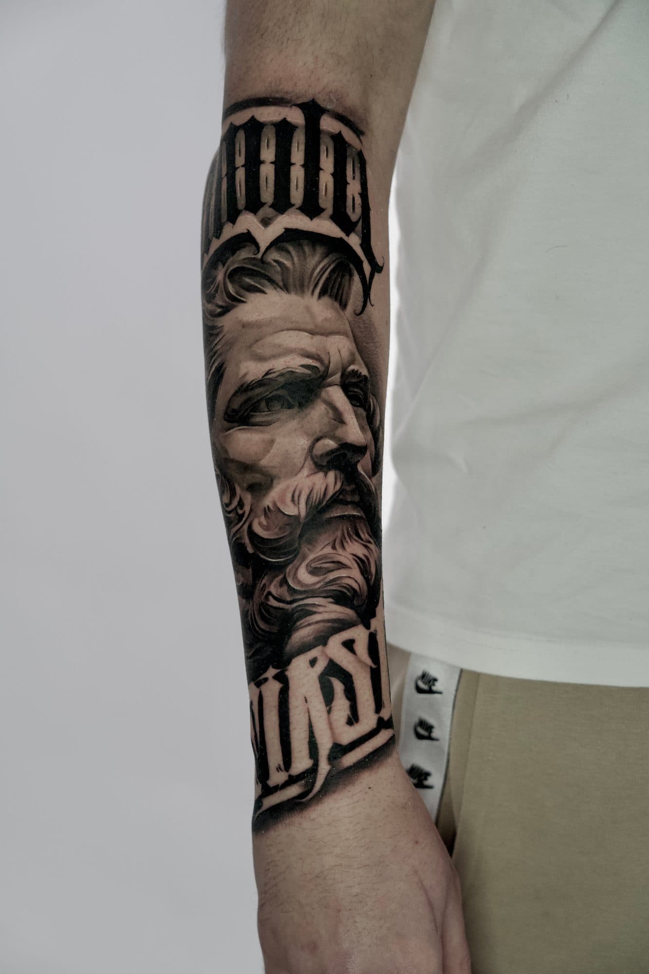 Antebraço em black and grey com retrato de figura mitológica barbada e lettering