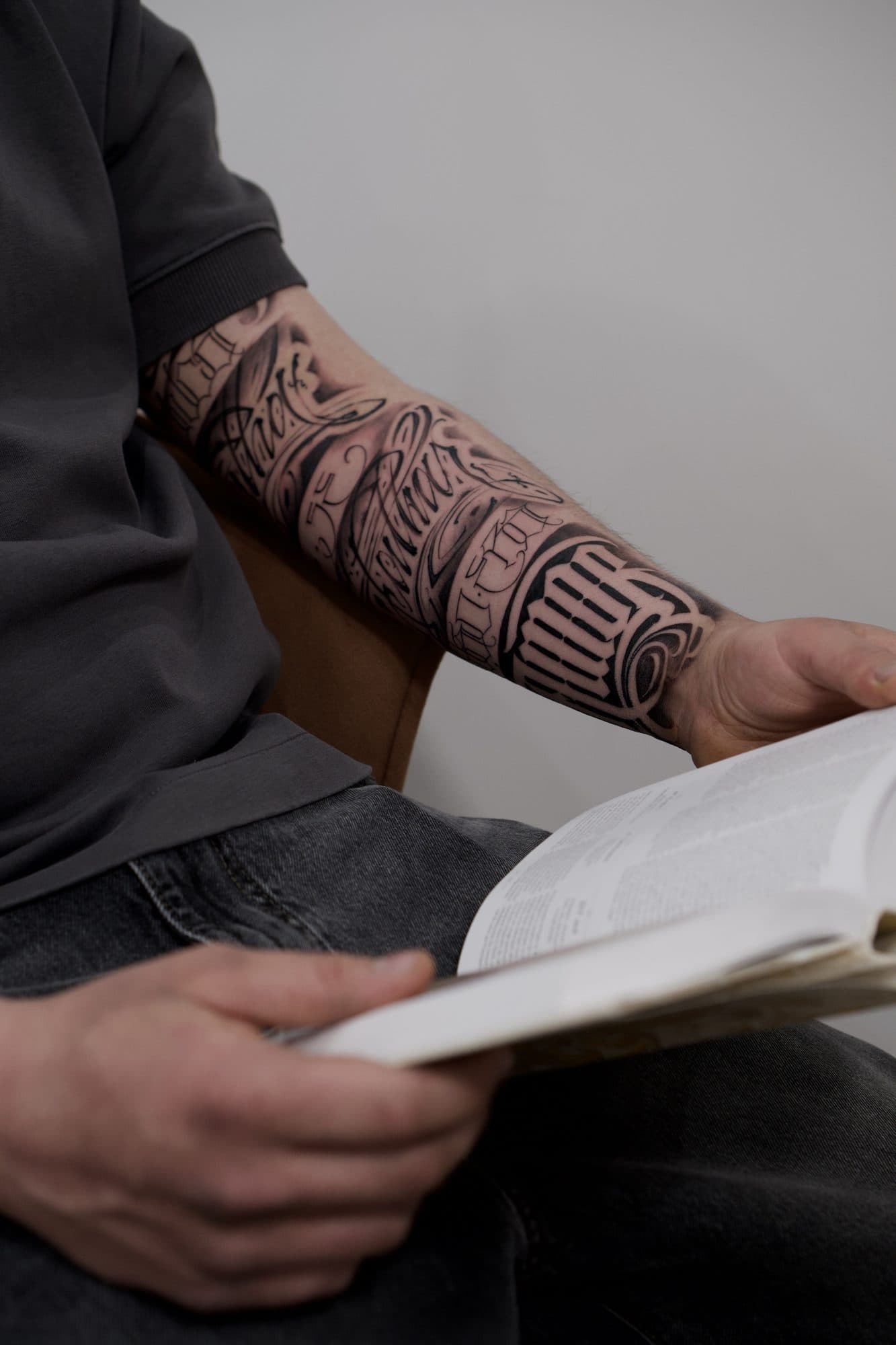 Manga de lettering no antebraço, com script e blackletter entrelaçados, num homem sentado a ler