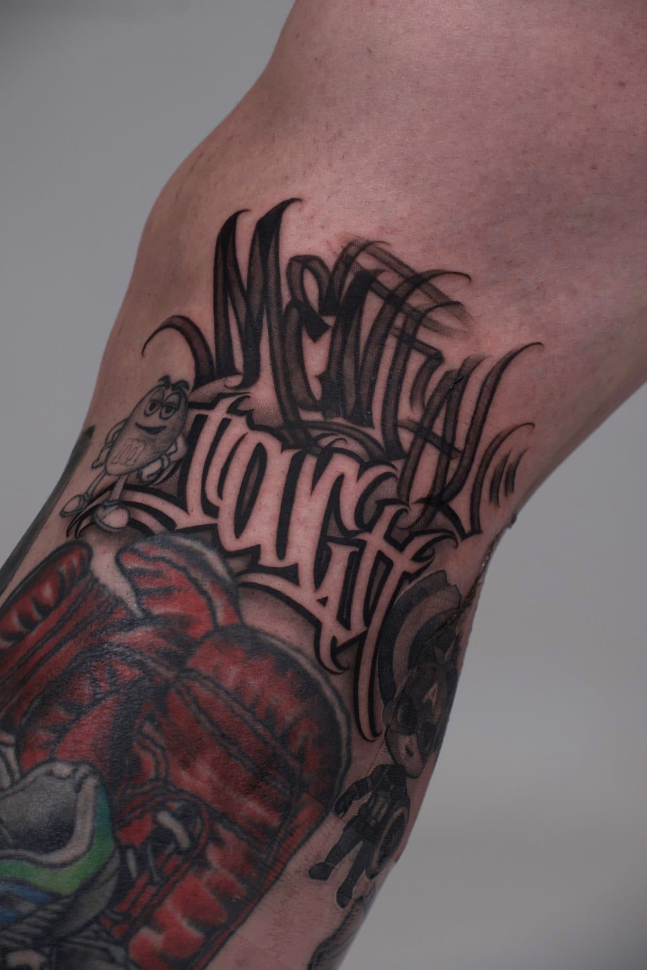 Lettering em blackletter no braço, por cima de uma tatuagem a cores